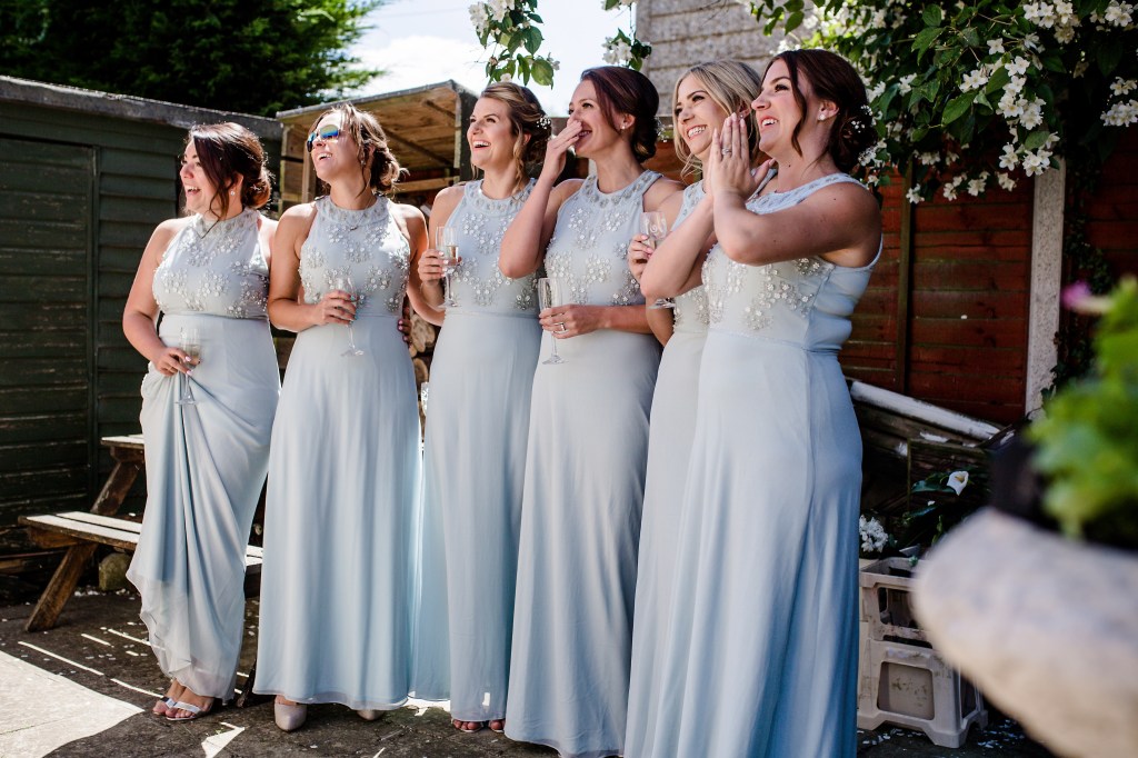 First look bridesmaids see te bride for the first time, this is called first look. Maidstone in sunny day, perfect day for a wedding at St Mary's church. In the photography background are white blossoming flowers that give this photographs nice and fresh touch.Bridesmaids are wearing blue neck dresses without sleeves and ont the front are crystals of white and blue clear colour.